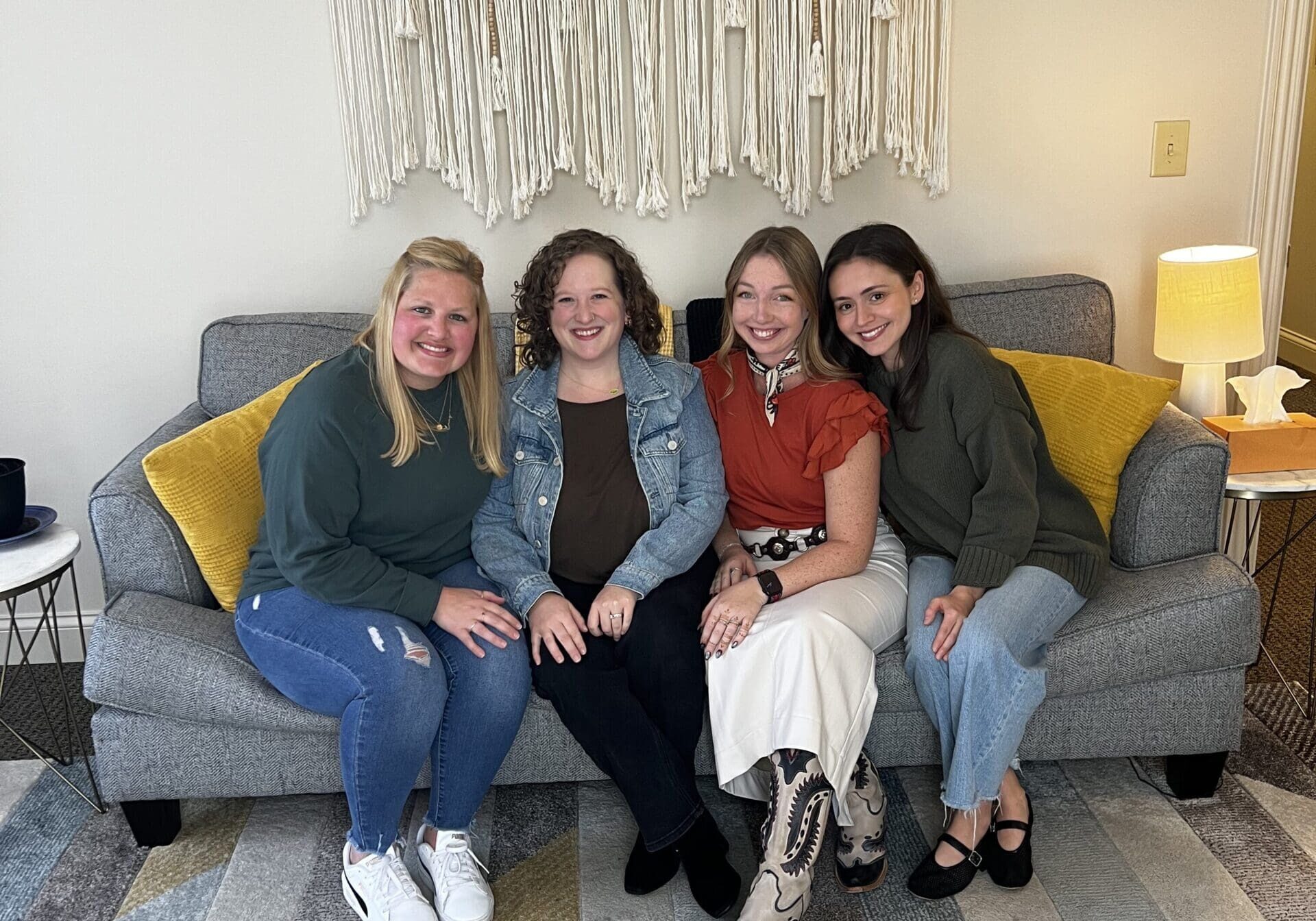 A group of four therapists sitting together on a gray couch in a cozy counseling office. They are smiling warmly, with a woven macramé wall hanging behind them and soft yellow pillows on either side.