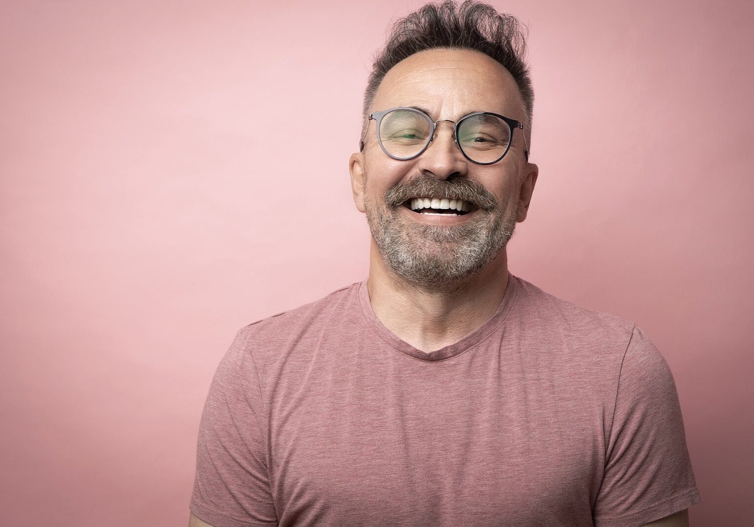 Middle-aged guy with trendy eyeglasses. Mature beard attractive man in t-shirt isolated on pink background. Portrait of happy expressive smiling 50s old man with mustache and glasses