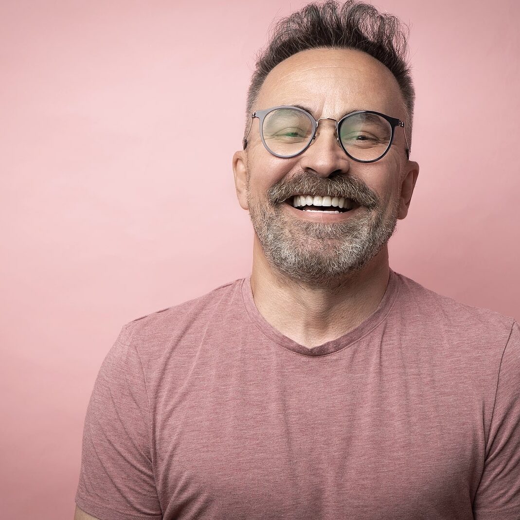 Middle-aged guy with trendy eyeglasses. Mature beard attractive man in t-shirt isolated on pink background. Portrait of happy expressive smiling 50s old man with mustache and glasses