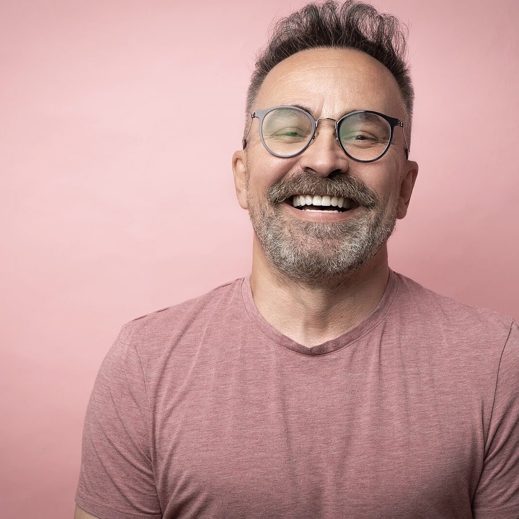 Middle-aged Guy With Trendy Eyeglasses. Mature Beard Attractive Middle-aged guy with trendy eyeglasses. Mature beard attractive man in t-shirt isolated on pink background. Portrait of happy expressive smiling 50s old man with mustache and glasses
