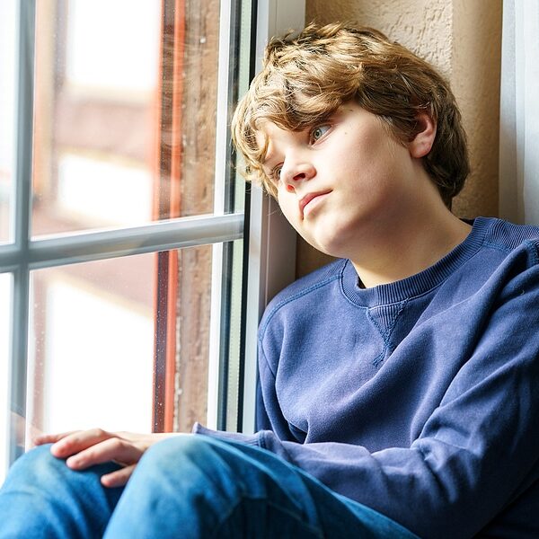 Sad pretten kid boy sitting on window. Unhappy angry child