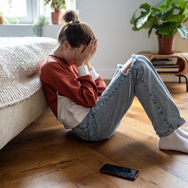 Unhappy teen girl covering face with hands and crying while sitting on floor with mobile phone nearby, upset frustrated child teenager being bullied or harassed online. Cyberbullying among teens