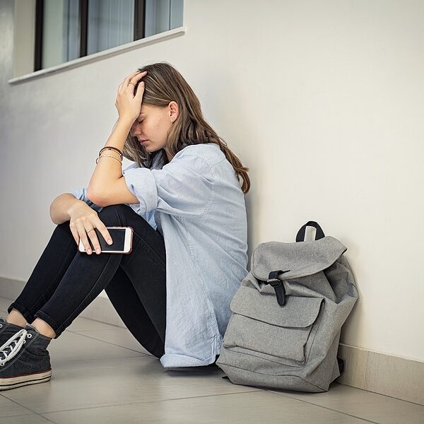 Upset and depressed girl holding smartphone sitting on college c Upset and depressed girl holding smartphone sitting on college campus floor holding head. University sad student suffering from depression sitting on floor at high school. Lonely bullied teen.